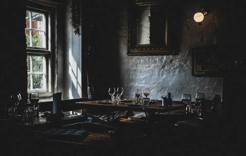 Laid tables against whitewashed stone walls in a dark dining room, a window at the back
