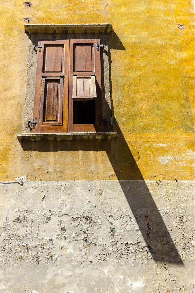 An ochre limewashed wall with a pair of wooden shutters half open in the afternoon sun
