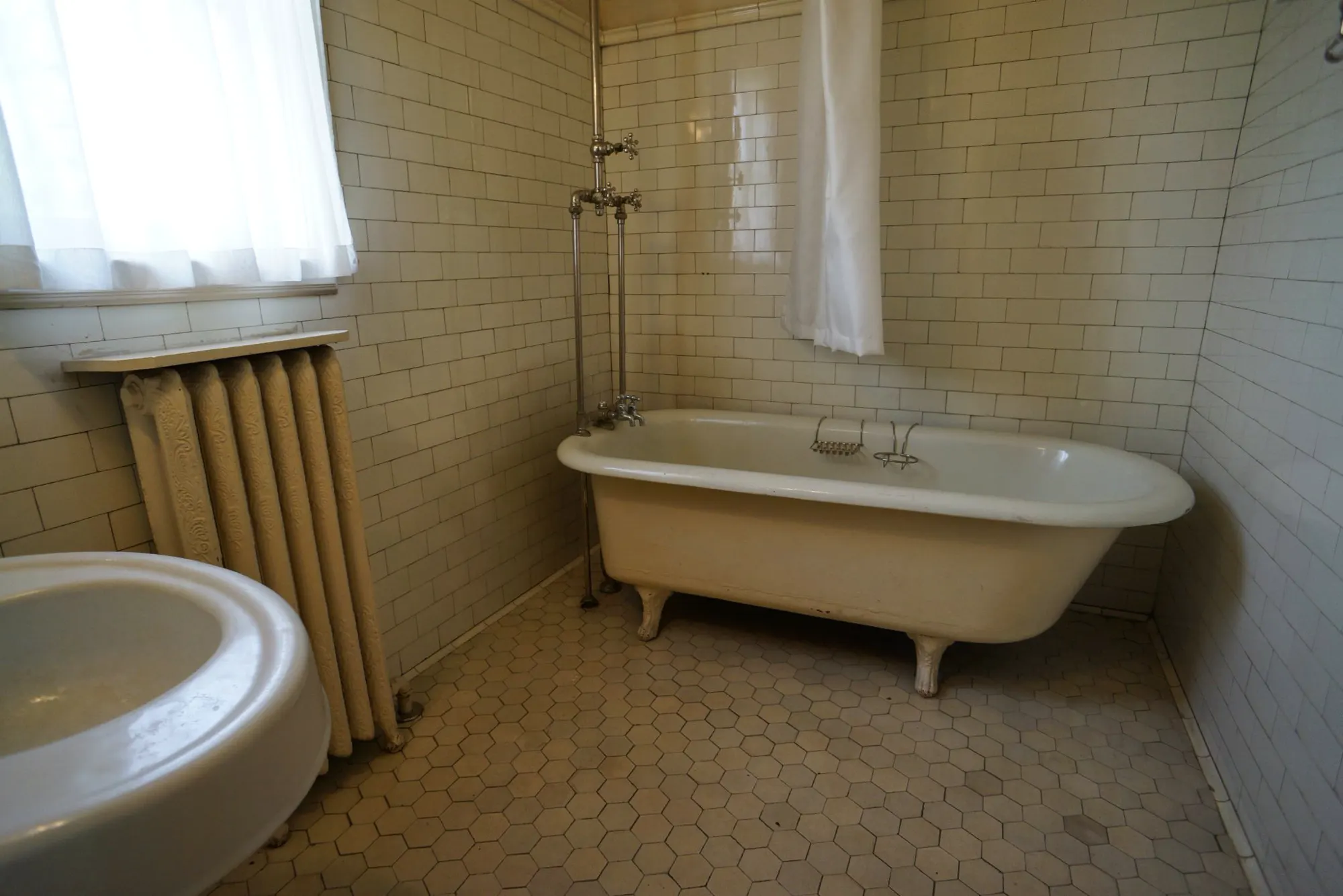 A clawfoot bath under a tall window in a white tiled bathroom